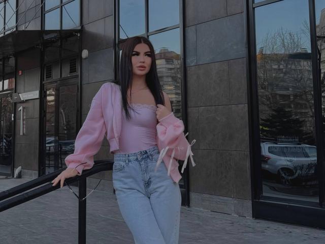 LeonaMeilee with long black hair, pink fluffy jacket, pink lace top, and high-waisted blue jeans, standing by a metal railing, hands in pockets, looking at camera.