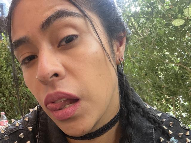 StellaBeckker's face, slightly open mouth, tongue partially out, pink lipstick, black braided hair, black choker, nose ring, black spiked jacket, green leafy background, brown eyes, medium skin.