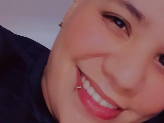 ScotTomboy's face, smiling with red lipstick, showing a silver lip piercing on the lower right side of their teeth. Soft, fair skin, slightly visible eyebrow, black shirt in the background.