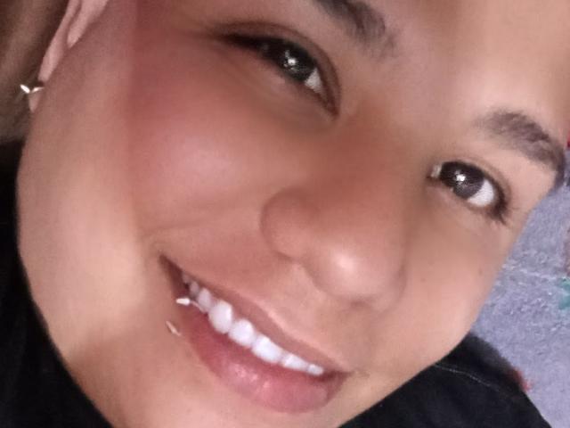 ScotTomboy's face with a smiling mouth, showing white teeth and a silver lip piercing. Light brown skin, dark eyes, and a pink headscarf with a small earring.