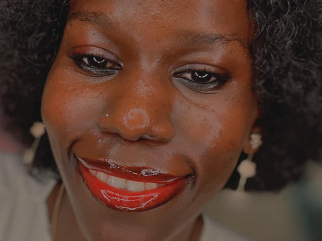 El rostro de KharlaQueen, piel marrón oscura, cabello negro rizado, lápiz labial rojo brillante, sonriendo, delineador de ojos, una pequeña marca de nacimiento sobre su nariz, aretes blancos, frente ligeramente brillante, sonriendo con labios rojos..