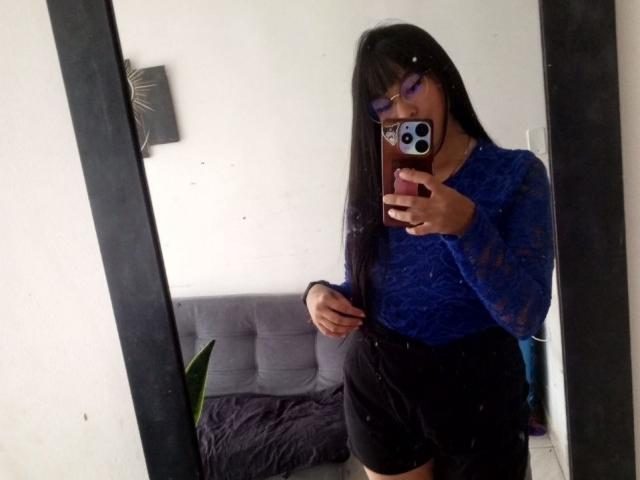 AndreaBrunert with long black hair, wearing purple glasses, blue lace top, black skirt, taking a mirror selfie, showing her small ass, holding a phone with a black case.