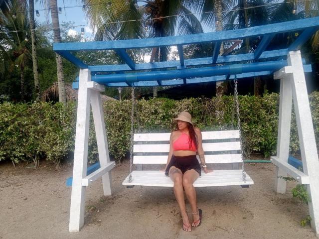 AndreaBrunert sits on a white wooden swing with blue overhead beams, wearing a pink crop top, black shorts, and a straw hat, in a tropical outdoor setting.