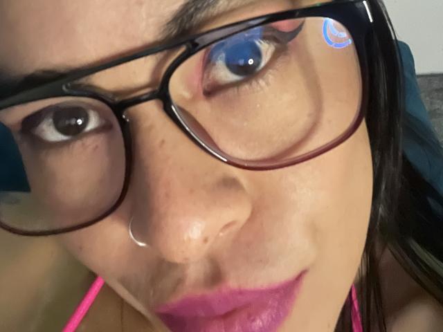 StellaBeckker's face with big black glasses, pink lipstick, nose ring, and a slight smile, showing her brown eyes and a small blue swirl sticker on the right glass.