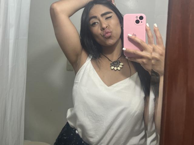 StellaBeckker, con un braccio alzato, labbra tumefatte, top bianco senza maniche, telefono rosa, collana nera e unghie bianche, sta scattando un selfie allo specchio, mostrando il seno..