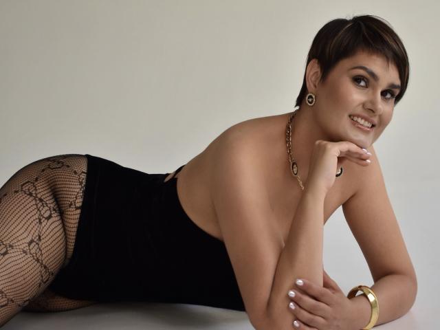 MayaNoir with short brown hair, smiling, laying on side in black lace-tightened dress, showing her ass, wearing gold necklace, bracelet, and earrings, with white-painted nails.