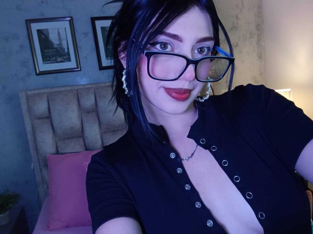 Isabella Rossi, con cabello negro, gafas y lápiz labial rojo, vestida con una camisa negra de botones que deja ver un escote pronunciado, mostrando sus senos, en un dormitorio con poca luz..