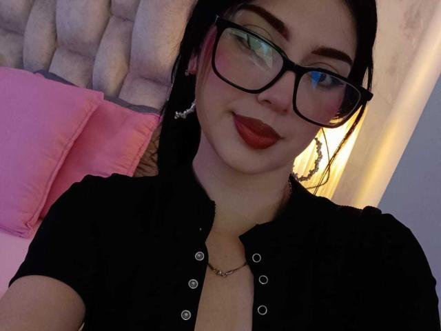 Isabella Rossi, con occhiali neri, rossetto rosso e camicia nera che mette in mostra il décolleté, sorride alla fotocamera, ha i capelli scuri, indossa orecchini e una collana. L'ambientazione è una camera da letto, con un cuscino rosa..