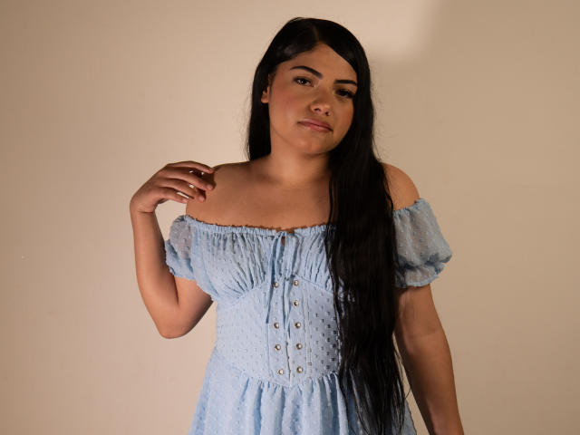 BrianaClarkX with long black hair, wearing a light blue off-shoulder dress, standing against a plain background, looking slightly to the side, hand near collar.
