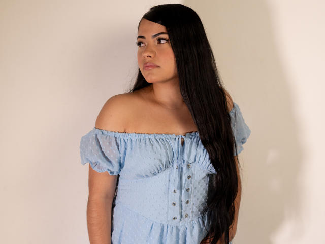 BrianaClarkX with long black hair, wearing an off-shoulder blue dress, buttoned down, showing her tits, standing against a white wall, looking to the side.