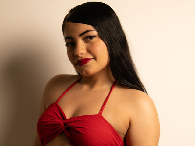 BrianaClarkX with long black hair, red lipstick, and a red halter top that ties at the center, showing her small tits and a cute smile.