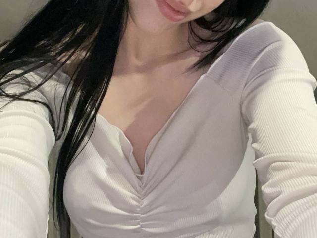 AmaiRin, con cabello largo y negro, vestida con una camiseta blanca de canalé, con puños y cuello en V, que muestra sus pequeños pechos firmes y un ligero escote, en un selfie..