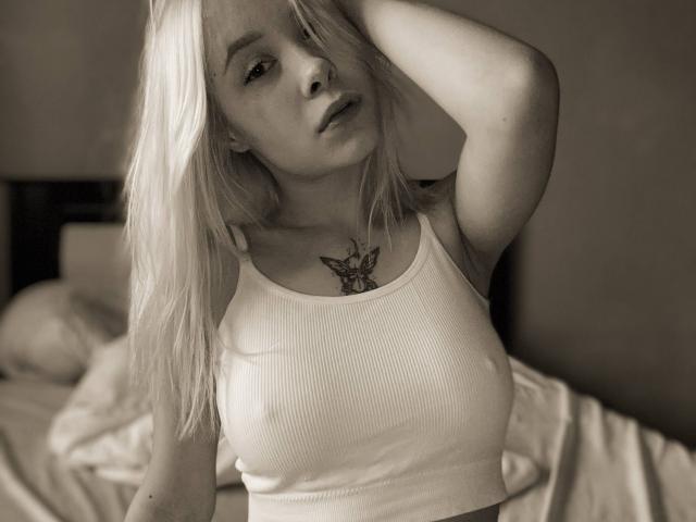 SoftAngelxKisses com cabelo loiro, vestindo uma camiseta branca texturizada, mostrando seios pequenos, braço levantado, tatuagem de borboleta no peito, com uma expressão ligeiramente entediada, em tons sépia, em uma cama..