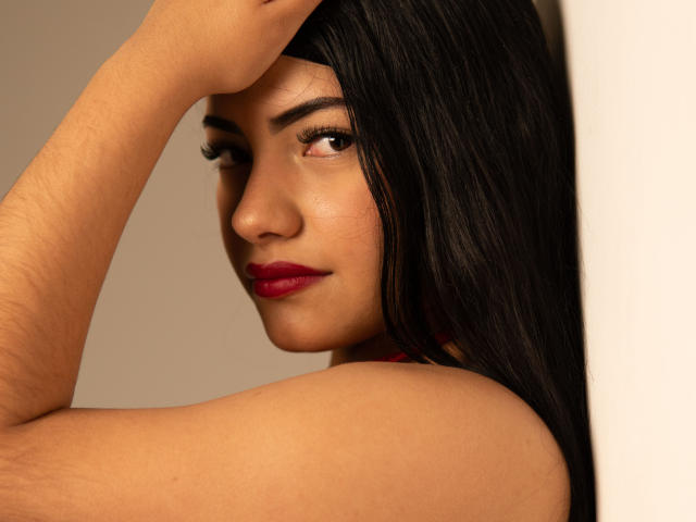 BrianaClarkX has tan skin, dark straight hair, red lipstick, and a slight smirk. She's raising her arm, showing her bare shoulder and subtle body hair.