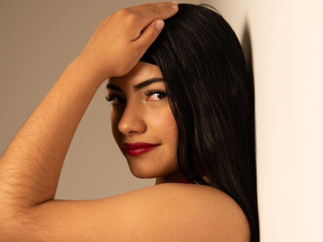 BrianaClarkX with dark hair, red lipstick, and brown eyes, holding her head with a hand, showing off her bare shoulders and a subtle smile. She's looking over her shoulder.