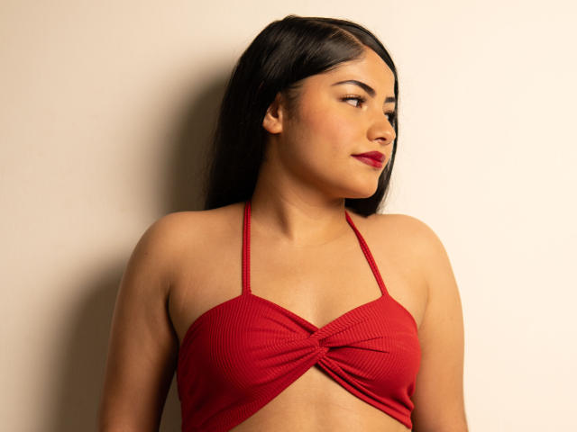 BrianaClarkX, com cabelo preto e liso, top vermelho de alças, mostrando seios pequenos, pele clara, batom vermelho, perfil lateral, olhando para a direita, em frente a uma parede bege lisa..