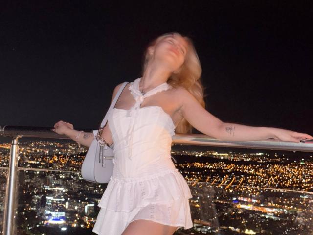 SoftAngelxKisses trägt ein weißes, gerafftes Kleid und lehnt sich nachts an ein Geländer, während die Lichter der Stadt im Hintergrund leuchten. Ihr Kopf ist leicht nach hinten geneigt, ihr blondes Haar fällt ihr herab. Sie hat kleine Tattoos am rechten Arm und trägt eine weiße Handtasche..