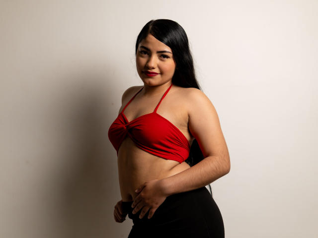 BrianaClarkX with long black hair, red bikini top showing her tits, black pants, and a slight smile, standing against a plain wall. Her hand rests on her hip.