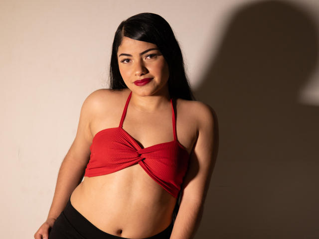 BrianaClarkX mit langen, schwarzen Haaren, einem roten, gerippten Crop-Top, kleinen Brüsten und schwarzen Hosen, steht vor einer schlichten Wand, mit einem Schatten auf der rechten Seite, und lächelt mit rotem Lippenstift..