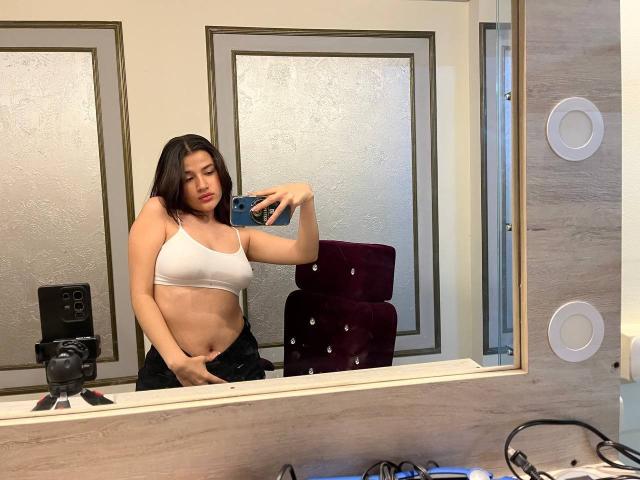 Iris Blair, com cabelo castanho, vestindo um top esportivo branco, expondo seus seios, segurando um telefone e cobrindo sua vulva com calças pretas em uma selfie no espelho do banheiro..