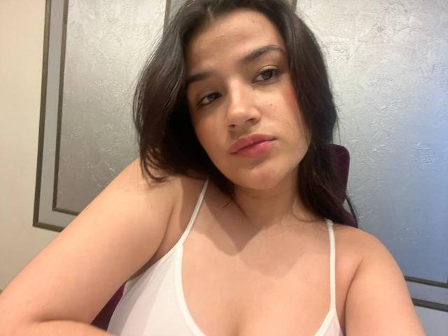 IrisBlair with dark brown hair, pale skin, and pink lips, wearing a white tank top, showing her small boobs, and a slight pout. Her arms are crossed.