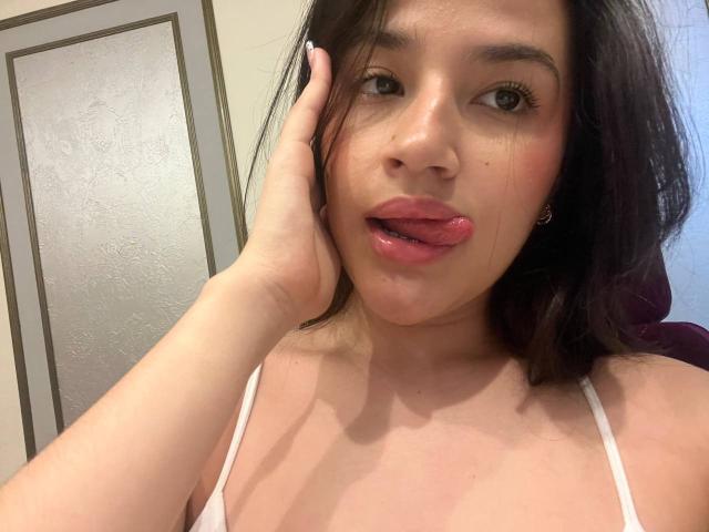 Iris Blair, met haar hand op haar wang, haar tong lichtjes uitgestoken, kijkend naar de camera, gekleed in een witte top, waarbij haar borsten zichtbaar zijn, met donkerbruin haar en subtiele make-up..