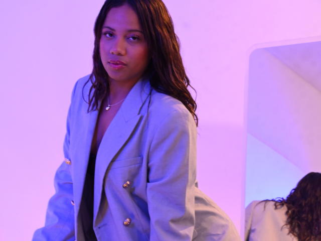 ZaraBlaze with dark brown wavy hair, wearing a light grey double-breasted jacket with gold buttons, looking slightly to the side, in a purple-lit setting.