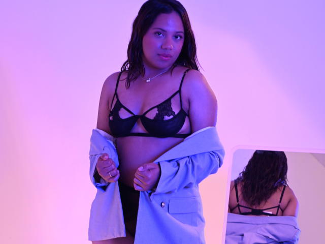 ZaraBlaze, brown-skinned woman with wavy black hair, wears black lace bra and gray jacket, showing her tits, mirrored reflection shows her back and bra straps. Purple-blue lighting.