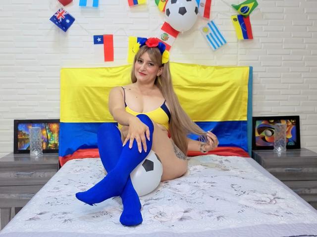 CristalMegan, con lunghi capelli biondi, indossa un reggiseno giallo e blu, calze blu abbinate, ed è seduta su un letto con un pallone da calcio, con il suo grande sedere e i suoi seni ben visibili..