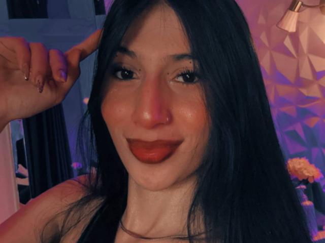 Sophie Beaumont tem cabelos escuros e lisos, lábios vermelhos cheios e um sorriso adorável, com uma mão tocando sua cabeça. Ela está em um quarto com pouca luz e iluminação roxa..
