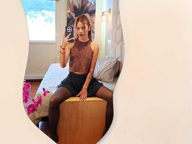 ValerieWoods, in a shiny brown sleeveless top and black shorts, sits on a wooden stool, wearing black stockings, taking a mirror selfie with a smartphone.