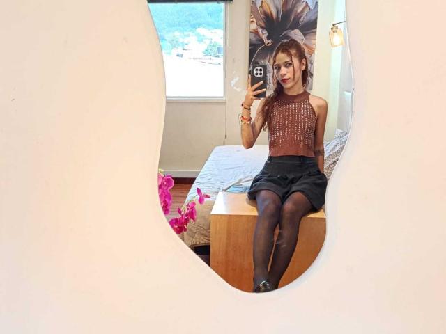  ValerieWoods with red lipstick, sitting on bed, wearing sparkly sleeveless top, black skirt, and black tights, holding phone, reflected in mirror, with pink flowers on left.