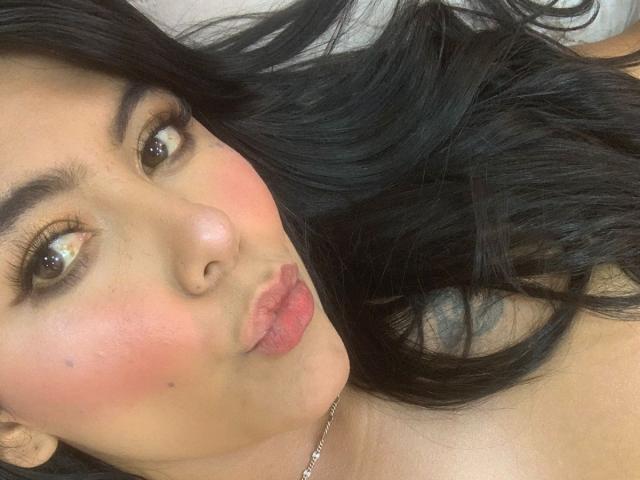 CatalinaReiyna with pouty lips, pink blush, black wavy hair, brown eyes, and a small beauty mark on her cheek, wearing a silver necklace, in a selfie.