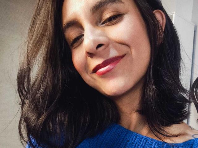 MiaAmoree, con cabello ondulado de castaño medio, sonriendo con lápiz labial rojo, una blusa de punto azul, la cabeza ligeramente inclinada y una expresión linda y segura. La luz natural y suave resalta sus rasgos..