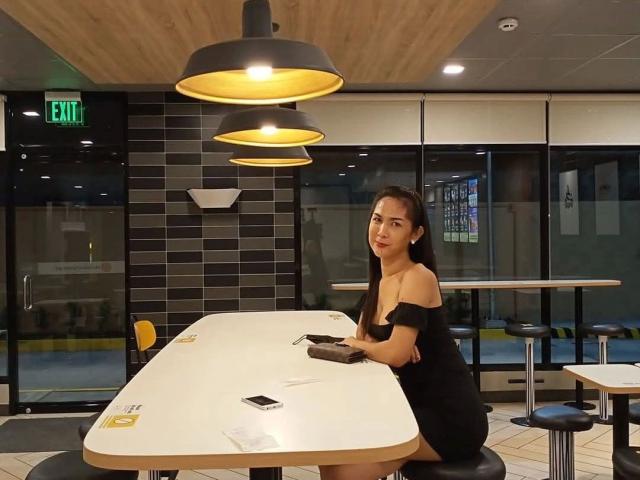 MISSKINKYWILD-hot with long dark hair, black off-shoulder dress, sitting at white table, hands on table, in modern cafe, under black and yellow hanging lights, with a phone.