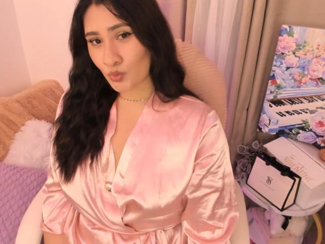 ElyRoss with wavy black hair, pouty lips, wearing a pink satin robe, sitting in a pink chair, looking at the camera, with a necklace. Boobs are not visible.