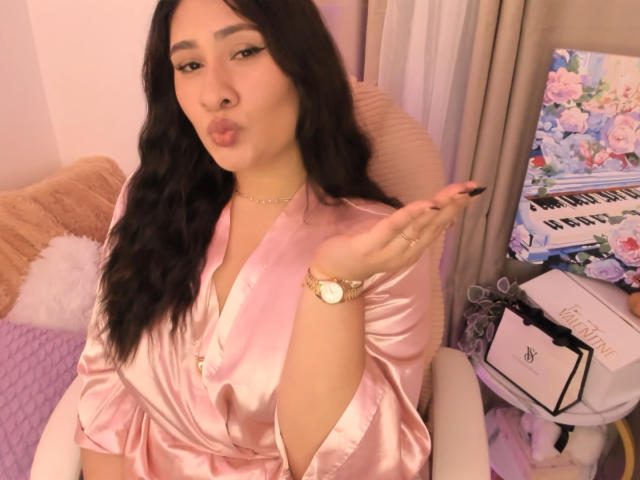 ElyRoss with long black hair, pouty lips, and pink satin robe, wearing a gold watch, sitting, hand up, black nails, pink pillow, beige chair, and floral keyboard.