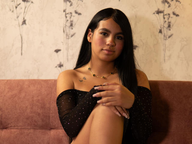 Dalila Tennesse, con lunghi capelli neri, indossa una maglia nera trasparente, seduta su un divano rosa, con le ginocchia sollevate, le mani sulle ginocchia, piccoli tatuaggi a forma di farfalla sul petto, una collana, un sorriso appena accennato..
