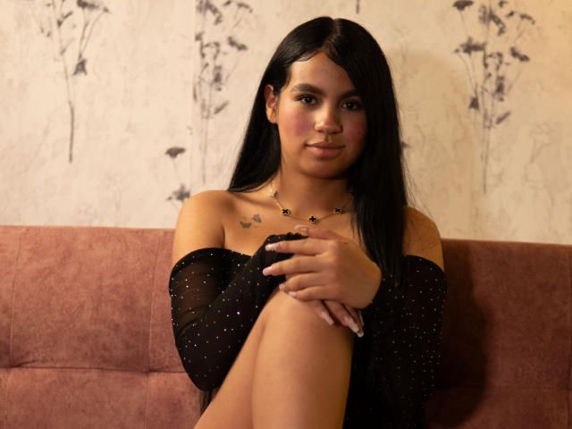 DalilaTennesse, with long black hair, sits on a pink couch, wearing a black off-shoulder top, her knees up, small butterfly tattoos on her chest, a subtle smile.