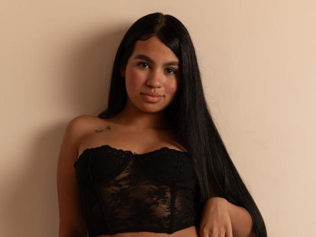 Dalila Tennesse, avec ses longs cheveux noirs, porte un haut noir en dentelle sans bretelles, a de petites seins et un tatouage sur son épaule droite, et elle sourit, debout devant un mur beige..