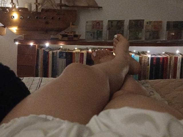 Las piernas de Genesiss, cruzadas a la altura de los tobillos, con las uñas de los pies pintadas de blanco, en una habitación con poca luz, vestida con pantalones cortos blancos, los pies apuntando hacia arriba, un modelo de barco de madera en el fondo..