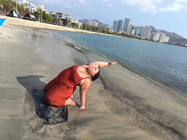HurrenMatt, chubby with big ass, wearing a red dress, kneels on wet sand, stretching back with arms, black hair, water and city skyline in background.