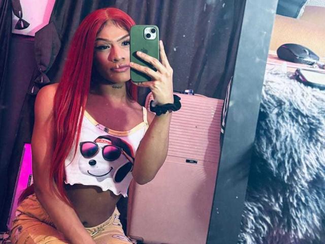 KeishaSexyBrood mit roten Haaren, kleinen Brüsten, trägt ein weißes Crop-Top mit einem Smiley und eine Sonnenbrille, hält ein grünes Telefon und sitzt vor einem Spiegel..