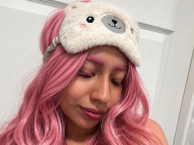 ZoeRedds, avec ses cheveux roses, portant un chapeau en forme d'ours blanc duveteux, a un eye-liner rose et un rouge à lèvres rose, les yeux fermés, souriant doucement, montrant son adorable nez et ses lèvres pulpeuses..