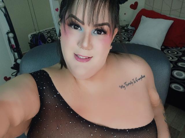 GiiannaMorrys with pale skin, black mesh top, large tits, tattoo on right shoulder, pink and blue eyeshadow, nose ring, smiling, brown hair, bed in background.