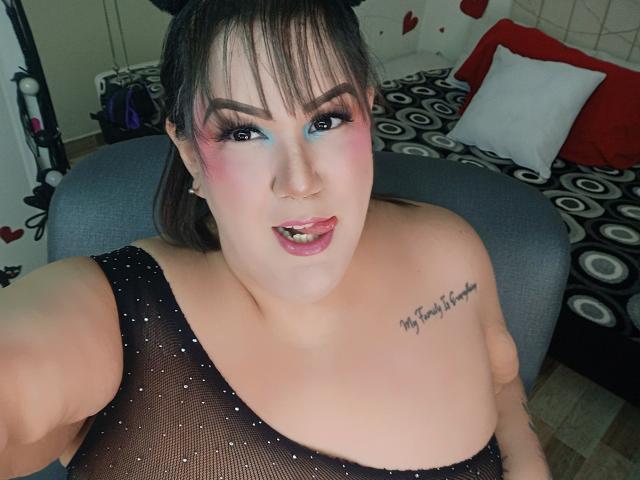 GiiannaMorrys, plus-sized with pale skin, dark hair in pigtails, wearing a sheer black top, showing her tits, tattoo on right shoulder, pink and blue eye makeup, tongue out.