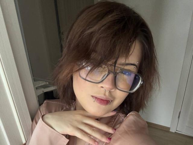 BarrieGromley with brown messy hair, glasses, nose ring, lip piercing, pink shirt, hand on chest, pink nails, looking at camera, light wood floor, white door in background.