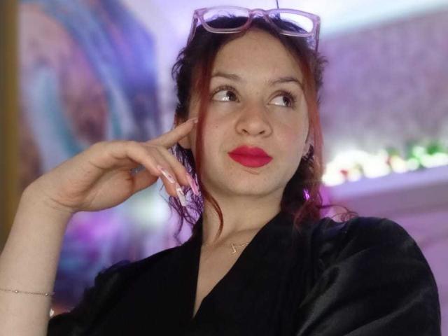 LesleyGray has red lipstick, black shirt, and glasses on head, looking up with hand near face, showing nails. Her ass is not visible. Her tits are not visible.