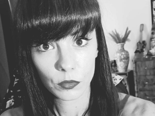 Black-and-white selfie of OrianaLaFrancaise with straight dark hair, bangs, and thick eyelashes. She has a pouty expression, wearing dark lipstick. Her face is slightly shiny.
