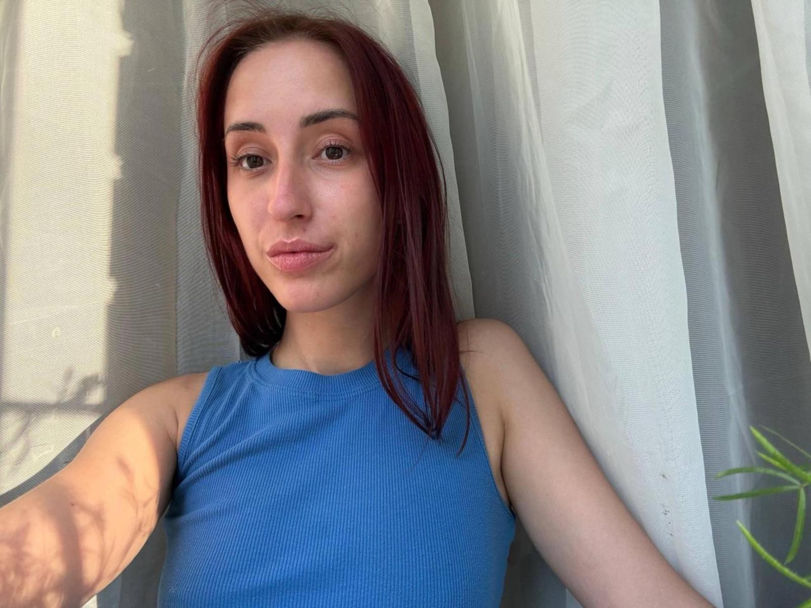 Vanita Rozenberg, con cabello rojo, vestida con una blusa azul sin mangas, con una ligera expresión de puchero, sentada frente a cortinas blancas, con la luz del sol proyectando sombras sobre su piel..